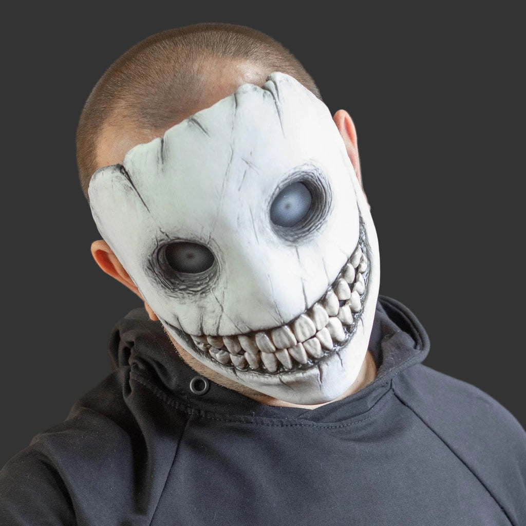 A person wearing a creepy, wide-grinning white mask with black cracks, hollow black eyes, and a sharp toothy grin, paired with a dark gray hoodie against a dark gray background.